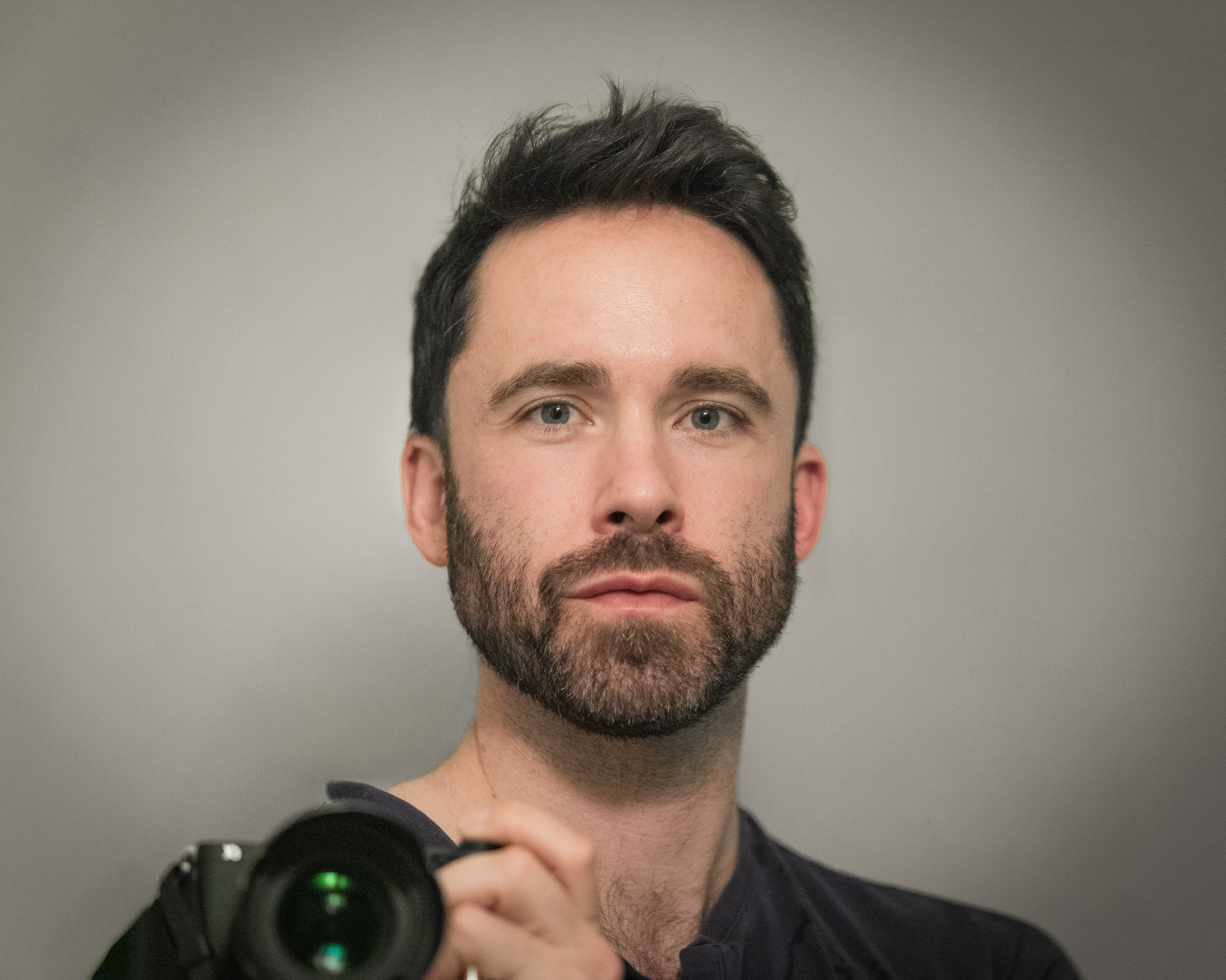 Color self-portrait of Yakov Barton holding a camera and looking directly forward.